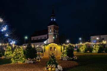 Festive Christmas lights, decorated trees, and a small holiday house glow warmly in front of a historic church on a winter night.