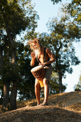 Man playing djembe drum outdoors on grassy hill surrounded by trees