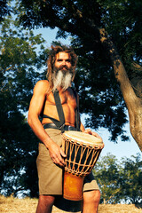 Intense bearded hippie drummer with djembe under tree in sunny forest