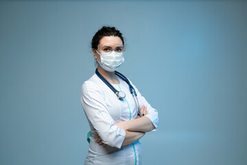 Medical worker in mask and protective glasses standing confidently with folded arms on blue background