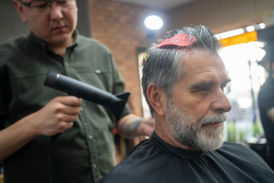 Barber drying hair of mature man with beard in professional barbershop