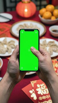 Vertical loop of a hand swiping on a green screen smartphone mockup, set against a festive Chinese New Year dinner background with red envelopes