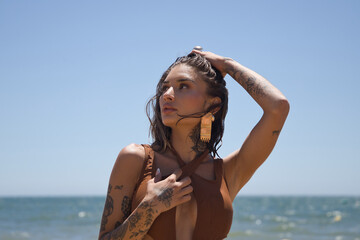 Portrait of a beautiful young Latina woman with tattoos wearing a modern brown swimsuit, with one hand on her wet hair, posing looking at camera, on the beach on a sunny day. Concept: vacation, summer