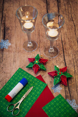 Christmas crafts at home: red and green 6-pointed paper stars with scissors and glue stick on a table in front of festive candlelight in vertical format.