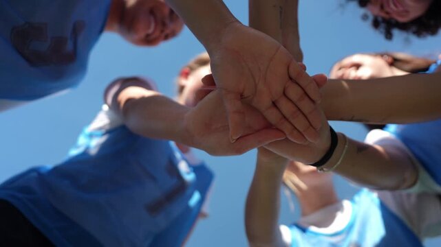 Low angle view of a women's soccer team putting their hands together in a huddle, then cheering and raising their arms to the sky