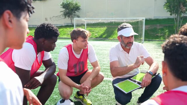 Professional male coach explaining game tactics on a clipboard to a diverse group of young soccer players squatting on the football field