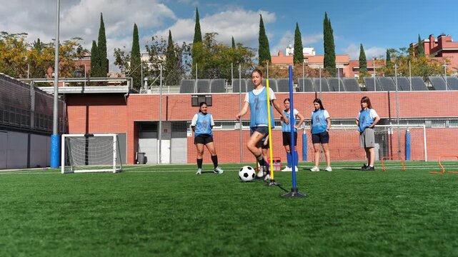 Group of female soccer players dribbling a ball around cones on a green turf field during a practice session on a sunny day - Powered by Adobe