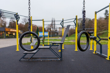Empty black fitness rings hang suspended at an outdoor public gym, featuring modern yellow and black exercise equipment for calisthenics training.