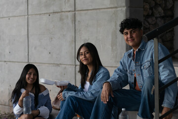 Group of young adult friends in casual denim clothes sitting on concrete steps, smiling while enjoying a break together and having a meal