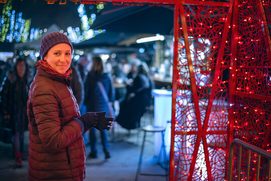 Close-up portrait of a cheerful woman (30s) bundled up in a winter hat and scarf, smiling happily at a cozy European Christmas market in Perpignan, France. The focus is on the warmth and seasonal joy