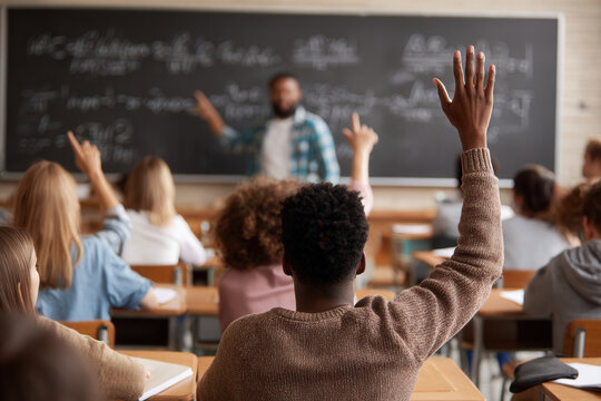 Students raise hands in classroom during lesson about mathematics or science with teacher explaining concepts at the blackboard