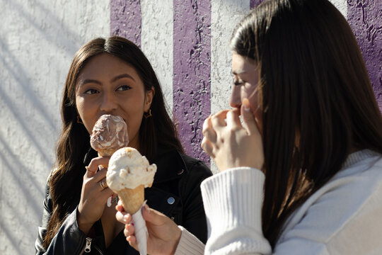 Two diverse young women enjoying ice cream cones together, sharing a sweet moment against a purple and white striped wall, celebrating friendship and a happy outdoor experience
