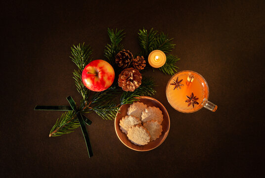 D&eacute;coration de No&euml;l, branche de sapin vert, pommes de pin, pomme, bougie en cire naturelle allum&eacute;e, petits g&acirc;teaux, tasse avec jus de pomme chaud, &eacute;pices de No&euml;l, vue du dessus, fond brun fonc&eacute;