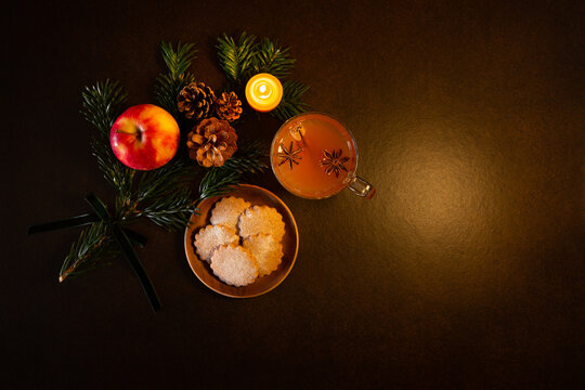Décor de Noël, branche de sapin vert, pomme, pommes de pin, bougie en cire naturelle allumée, petits gâteaux, tasse en verre avec jus de pomme chaud et épices de Noël, vue du dessus, fond brun foncé