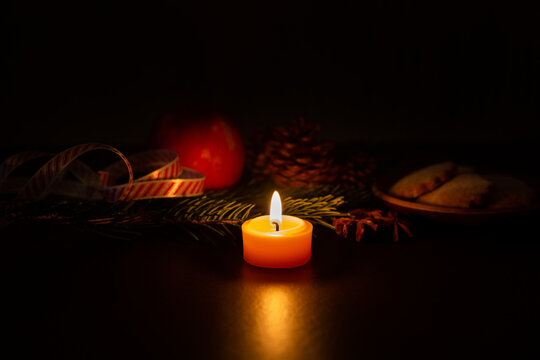 Petite bougie en cire naturelle orange allum&eacute;e avec une flamme centr&eacute;e dans l&rsquo;image, ambiance de No&euml;l, branche de sapin, ruban satin rouge et blanc, pomme, pomme de pin, petits g&acirc;teaux et anis &eacute;toil&eacute;e