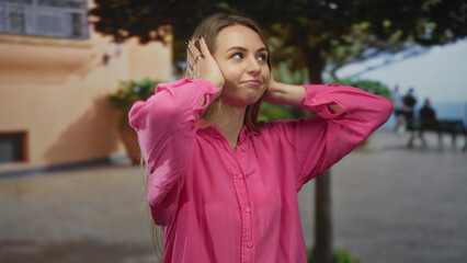 Young woman in pink shirt outdoors covers ears with hands, conveying a sense of peace amid urban scenery, her serene expression adds to the tranquil atmosphere.