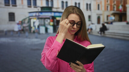 Woman reading book in urban street setting with blurred city background wearing glasses pink shirt...
