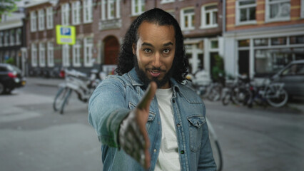 Man extends hand for handshake on street in city, denim jacket visible and bicycle row behind him;...