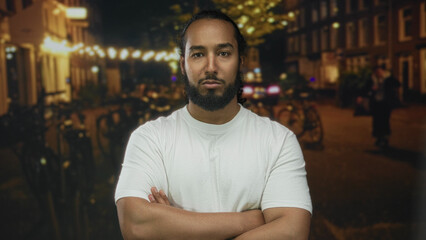 Man standing with arms crossed showing forearms and beard on a city street at night, wearing white...