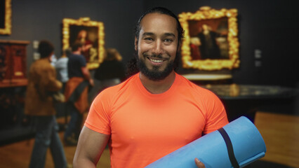 Man holding blue yoga mat and smiling while standing near framed paintings in gallery building wearing bright orange shirt; serenity curiosity.