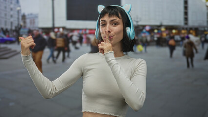 Young woman wearing cat ear headphones places finger to lips for silence amid blurred street crowds; mystery.