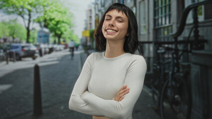 Fototapeta premium Woman wearing white crop top stands smiling with arms crossed on a bustling city street lined with parked bicycles; confidence.