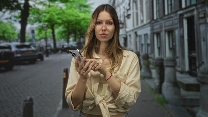 Fototapeta premium Woman tapping smartphone with finger on screen while standing on a cobbled street near parked cars and historic row buildings; focused connection.