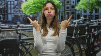 Woman points both index fingers upward on amsterdam canal street, puffing cheeks and smiling while gesturing to the sky; playful travel memory.