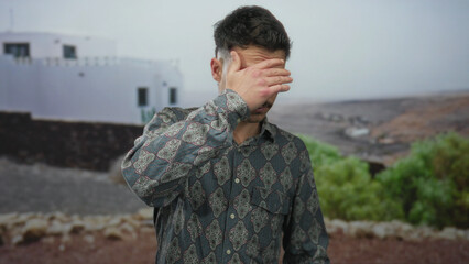 Young man covers face standing outdoors with a patterned shirt against a rural backdrop featuring plants and a distant building suggesting an introspective moment.