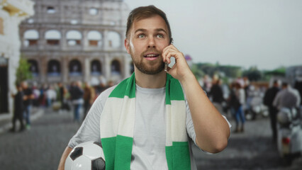 Man holding soccer ball and phone to ear in front of coliseum building, wearing green scarf and...