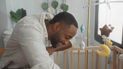 Young man smiling and leaning over wooden crib while playing with plush mobile in baby nursery; tenderness bonding.