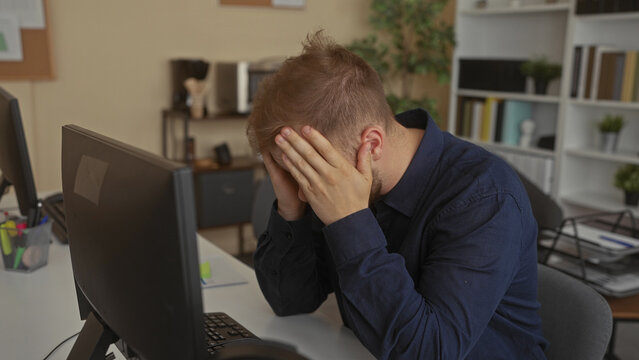 Man with hands on cheeks removing glasses while staring at computer monitor at desk inside building; stress concentration.