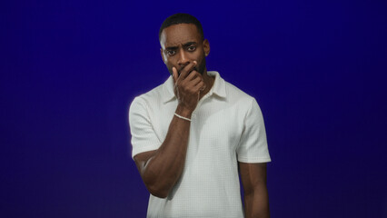 Man with hand covering mouth and visible beard and bracelet in studio against deep blue backdrop; concern thought.