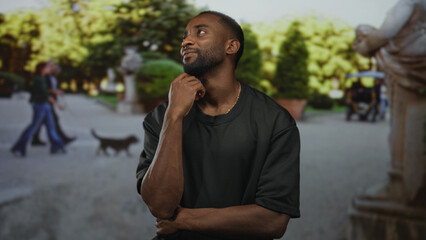 Young african american man with hand on chin, thinking pose on street by park statues and trees,...