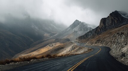 A winding road stretches through a rugged mountain range with rocky peaks and patches of grass. Clouds hover above creating a dramatic atmosphere during the afternoon.