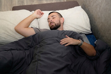 A man lies in bed under a blanket, appearing to sleep. His face shows signs of fatigue. It seems he is taking time off for recovery at home during the daytime