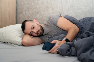 A man lies in bed under a blanket, checking his phone. He looks tired and is on sick leave to recover from an illness. The time is likely during the day