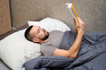 A man lies in bed with a blanket, reading a book. He shows signs of fatigue as he takes time to recover from an illness while resting at home