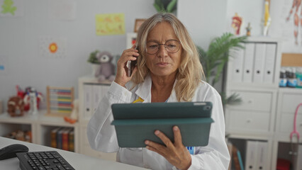 Woman pediatrician holding tablet and phone to ear in building office with stethoscope visible and children's toys on shelves; telehealth consultation compassion.
