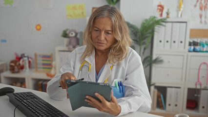 Woman pediatrician in white coat points pen at tablet on clinic desk with keyboard, binders and toys visible; calm compassion care.