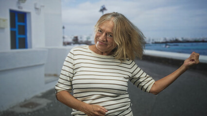 Fototapeta premium Senior woman with raised hand and open arm gesture laughing on a coastal street beside white building and seawall, wearing striped shirt; joy.