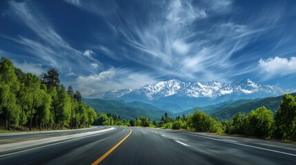 A highway stretches through verdant hills surrounded by trees and stunning snow covered mountains. The bright blue sky features wispy clouds creating a picturesque driving experience.
