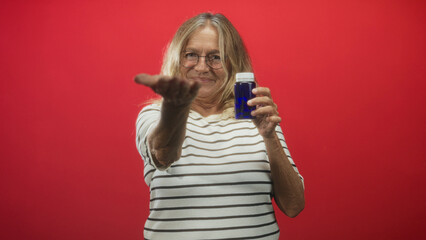 Senior woman holds blue pill bottle in right hand and extends left palm forward while smiling in studio; health reassurance.