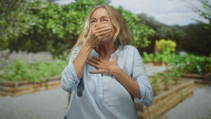 Senior woman covers mouth with hand and clutches chest for heartattack among raised planters in a...