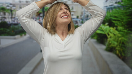 Fototapeta premium Middle aged woman in white shirt shades eyes with hands on an urban street under bright sunlight; optimism.