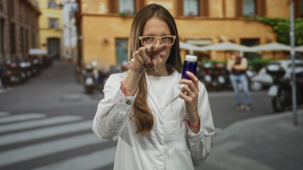 Young hispanic scientist woman in white lab coat points finger at camera on city street while holding a small blue bottle; health warning.