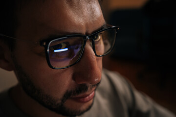 Close-up face of serious software developer wearing glasses, focused looking on computer screen reflecting in lenses while working late into night in dark office environment.