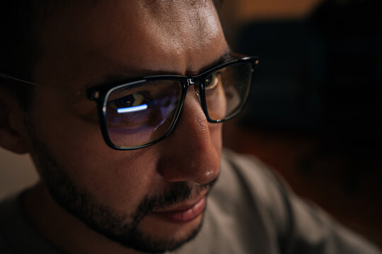 Close-up face of serious software developer wearing glasses, focused looking on computer screen reflecting in lenses while working late into night in dark office environment.