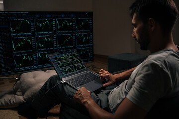 Back view of successful trader engrossed in financial analysis and day trading, using laptop and large external monitor displaying various stock market and cryptocurrency charts from home office.