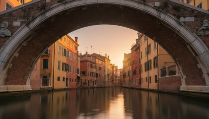 Naklejka premium Picturesque European canal with colorful buildings and calm water reflections is framed by an old stone bridge arch during a beautiful golden hour sunset
